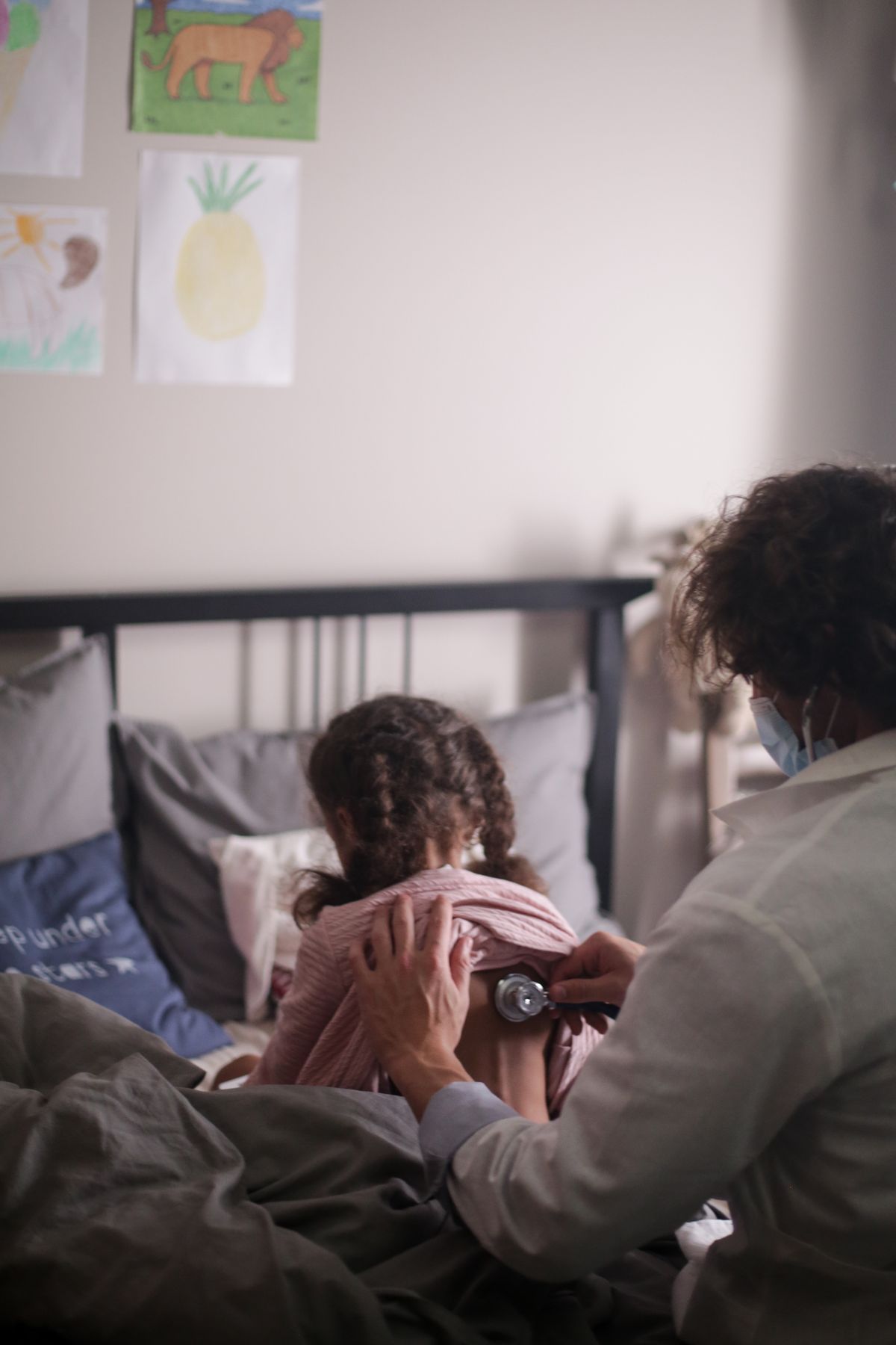 Un médecin ausculte une fillette assise sur son lit, dans sa chambre.